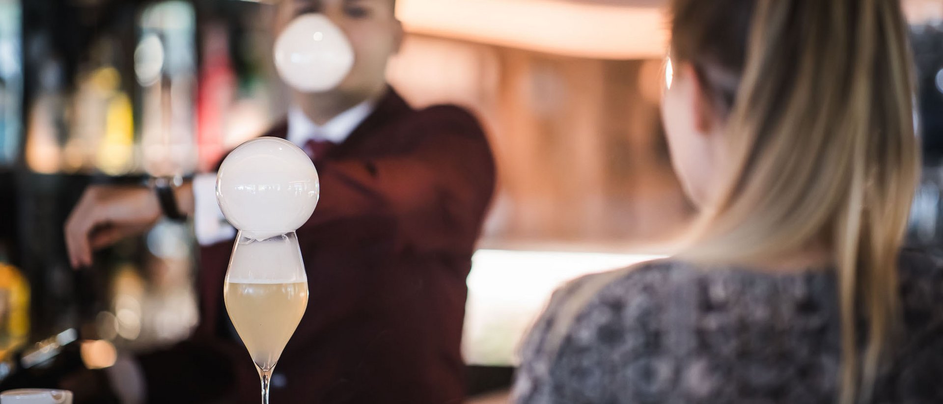 Bartender prepara cocktail con bolla di schiuma, donna seduta al bancone