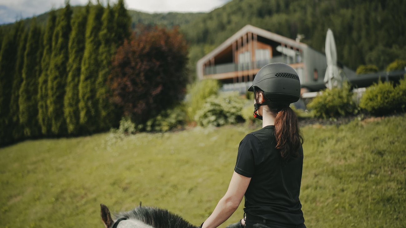 Außergewöhnliche Erlebnisse, Reiten und Yoga im Urlaub Frau mit Helm reitet Pferd vor Haus in grüner Landschaft