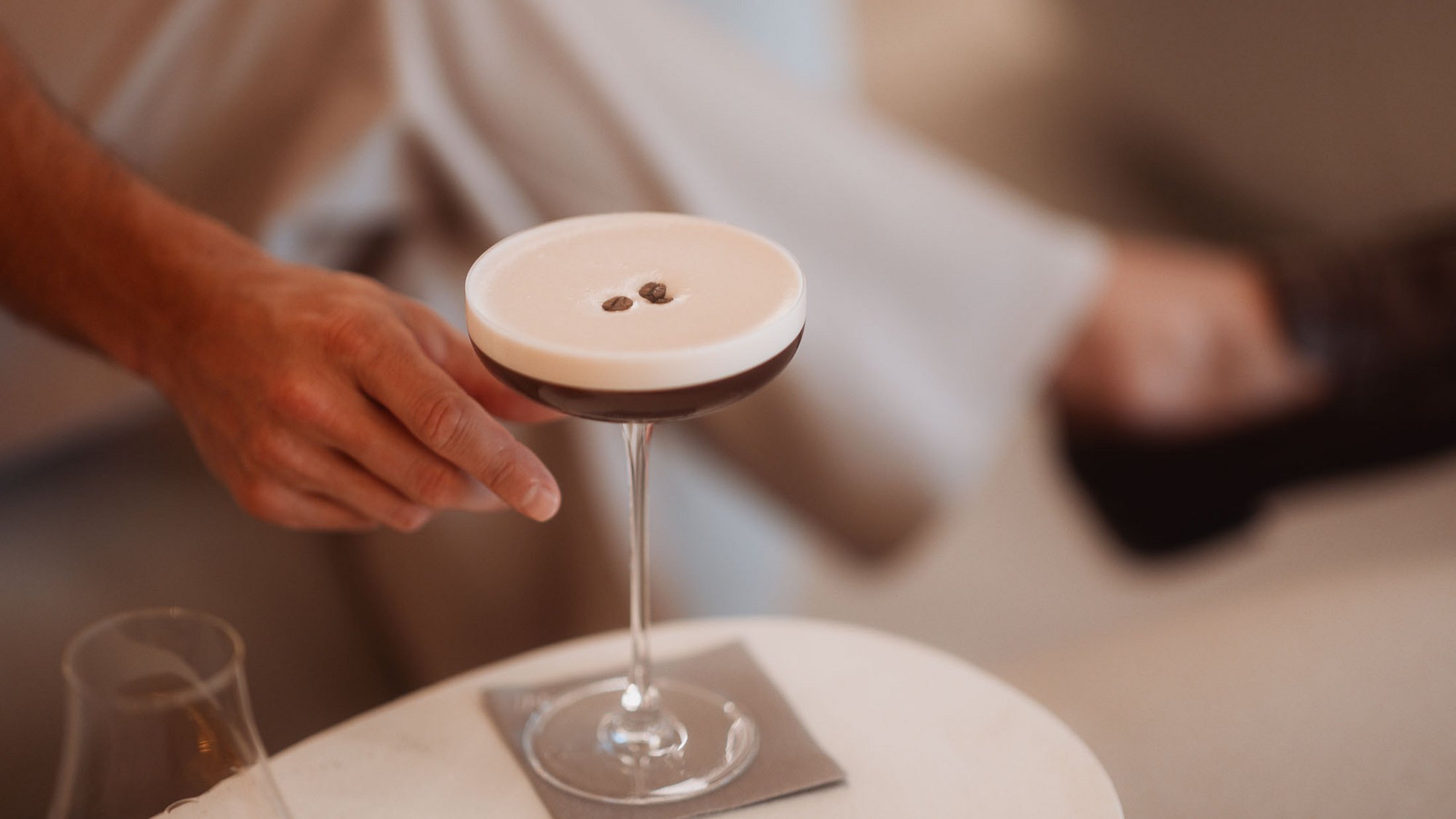 Mano che tiene un cocktail con schiuma e chicchi di caffè su piccolo tavolo bianco