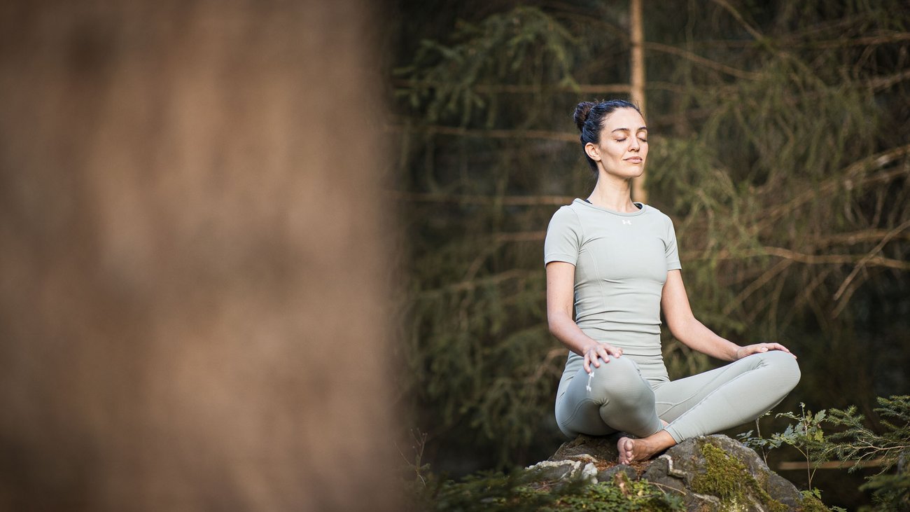 Frau meditiert im Yoga-Sitz auf moosbedecktem Felsen im Wald
