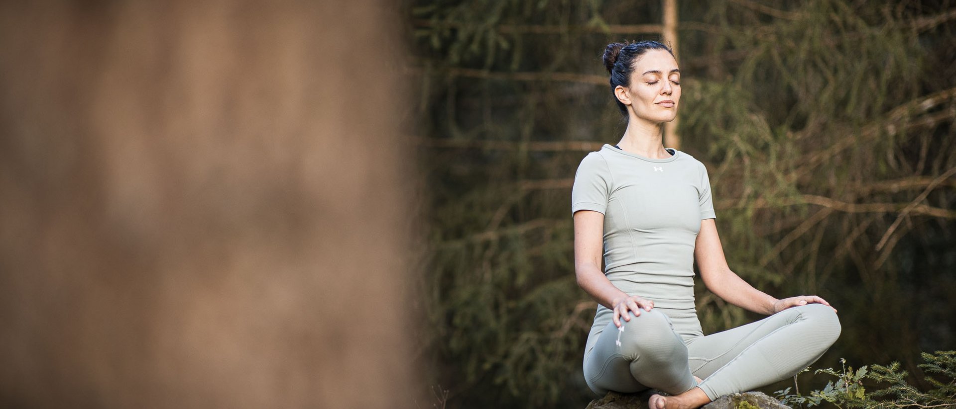 Frau meditiert im Yoga-Sitz auf moosbedecktem Felsen im Wald