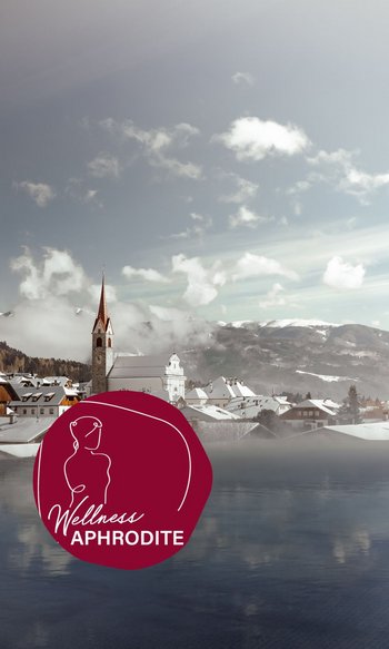 Die Heimat des Strahlens: Ihr Wellnesshotel in Südtirol Verschneites Dorf mit Kirchturm vor Bergen und See, mit Wellness-Aphrodite-Logo