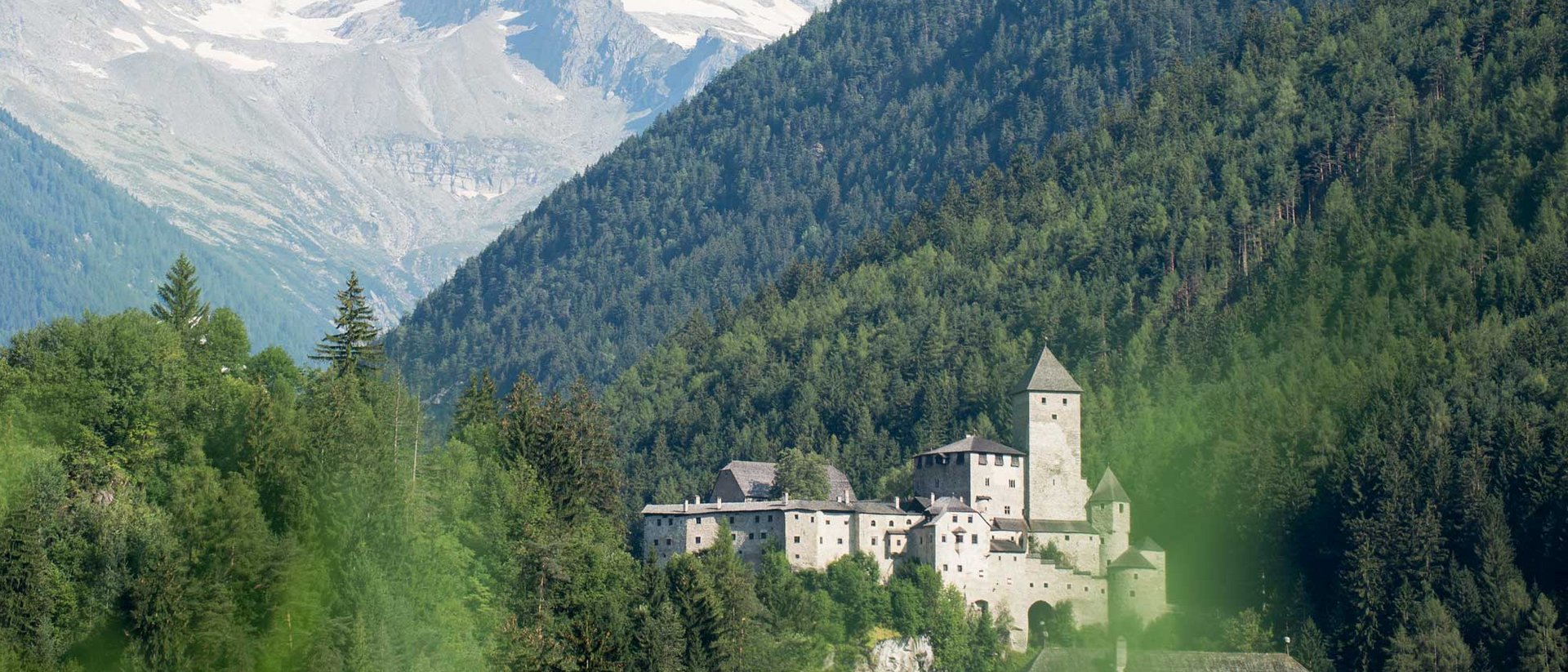Burg in den Alpen mit bewaldeten Hügeln und schneebedeckten Bergen im Hintergrund
