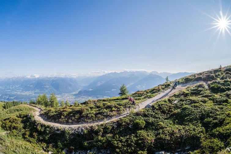Two mountain bikers on a sunny trail with mountains and clear sky in the background
