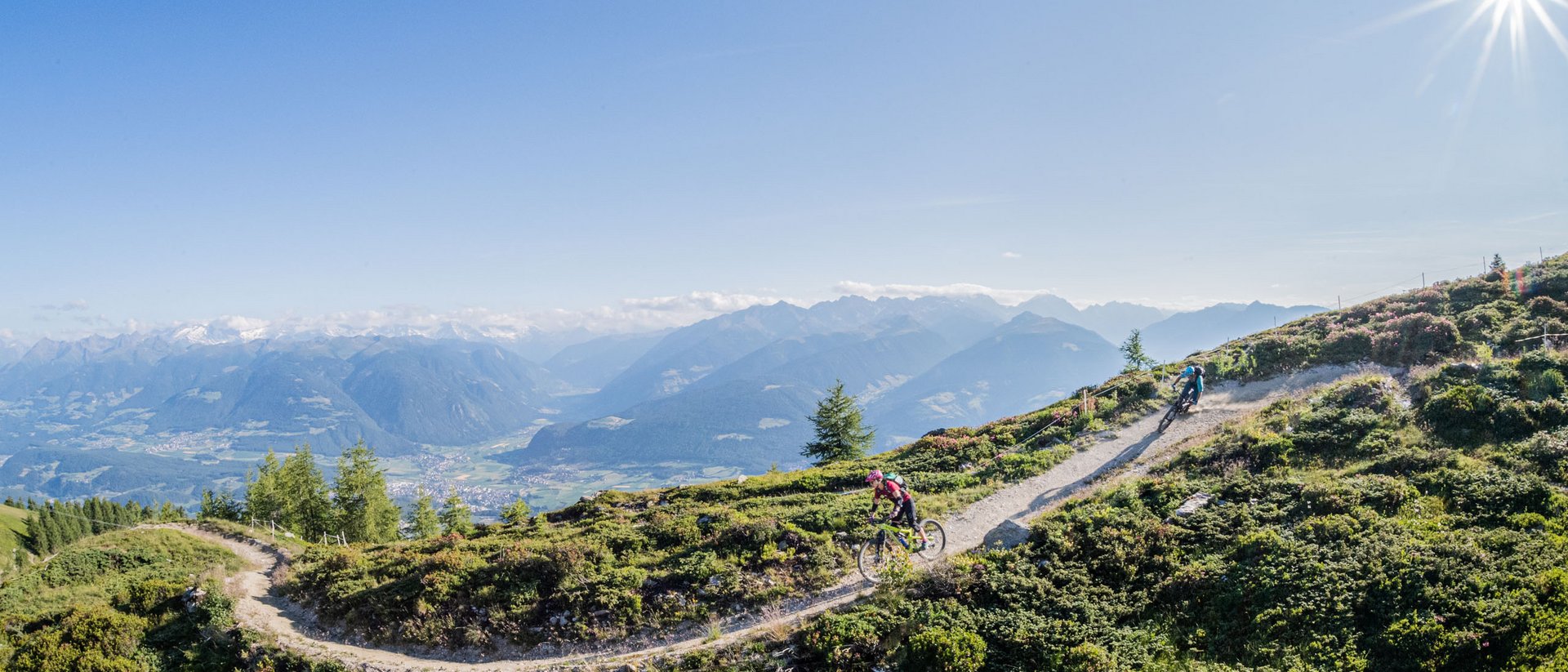 Zwei Mountainbiker fahren auf einem Bergweg mit sonnigem Himmel und Alpen im Hintergrund
