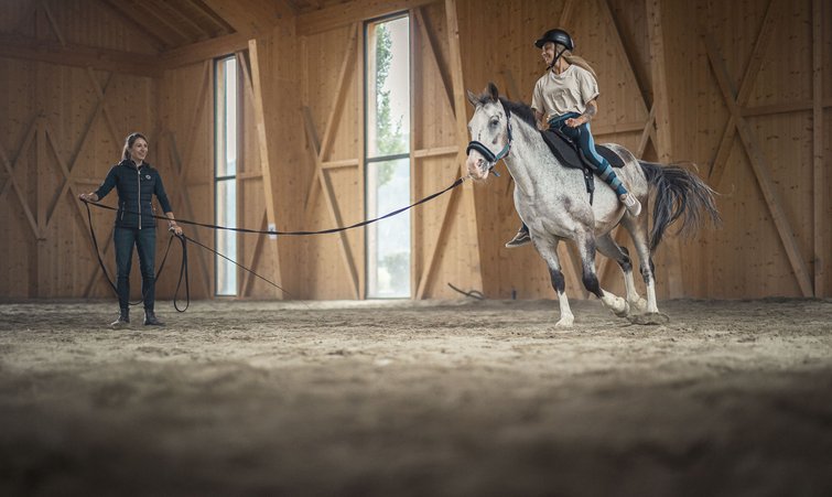 Woman riding horse in indoor arena with trainer holding longe line