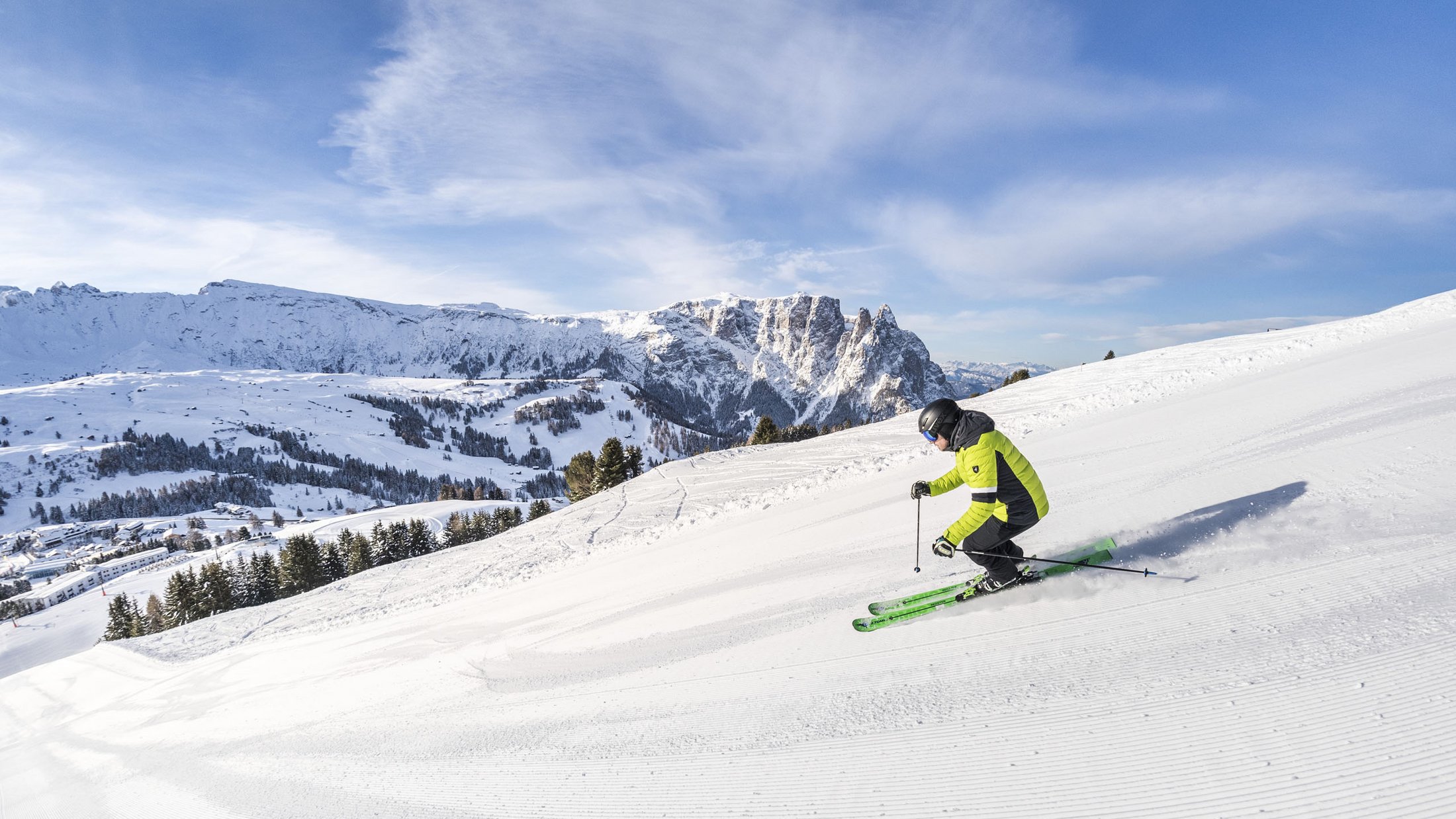 Sciatore con giacca verde su pista battuta con montagne sullo sfondo