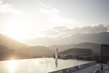 Eine regenerierende Auszeit vor Weihnachten Person im Bademantel am Infinity-Pool bei Sonnenuntergang mit Bergblick