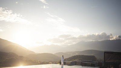 Persona in accappatoio alla piscina a sfioro al tramonto con vista sulle montagne