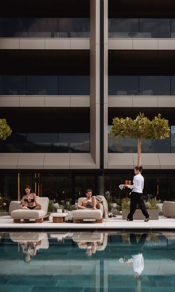 Zwei Menschen entspannen auf Liegestühlen am Pool, Kellnerin bringt Getränke