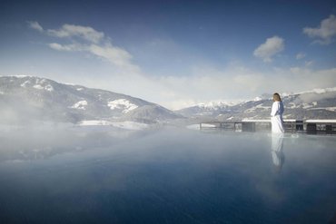 Frau in Bademantel am beheizten Außenpool mit Blick auf schneebedeckte Berge