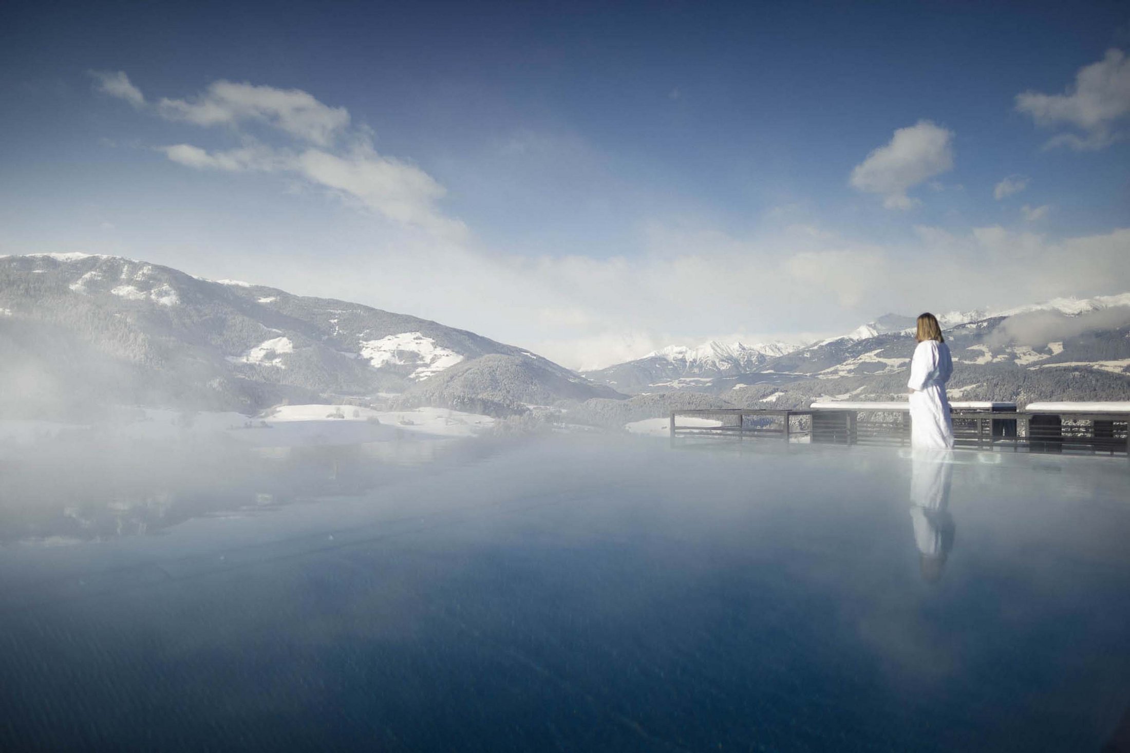 Woman in bathrobe by heated outdoor pool overlooking snowy mountains