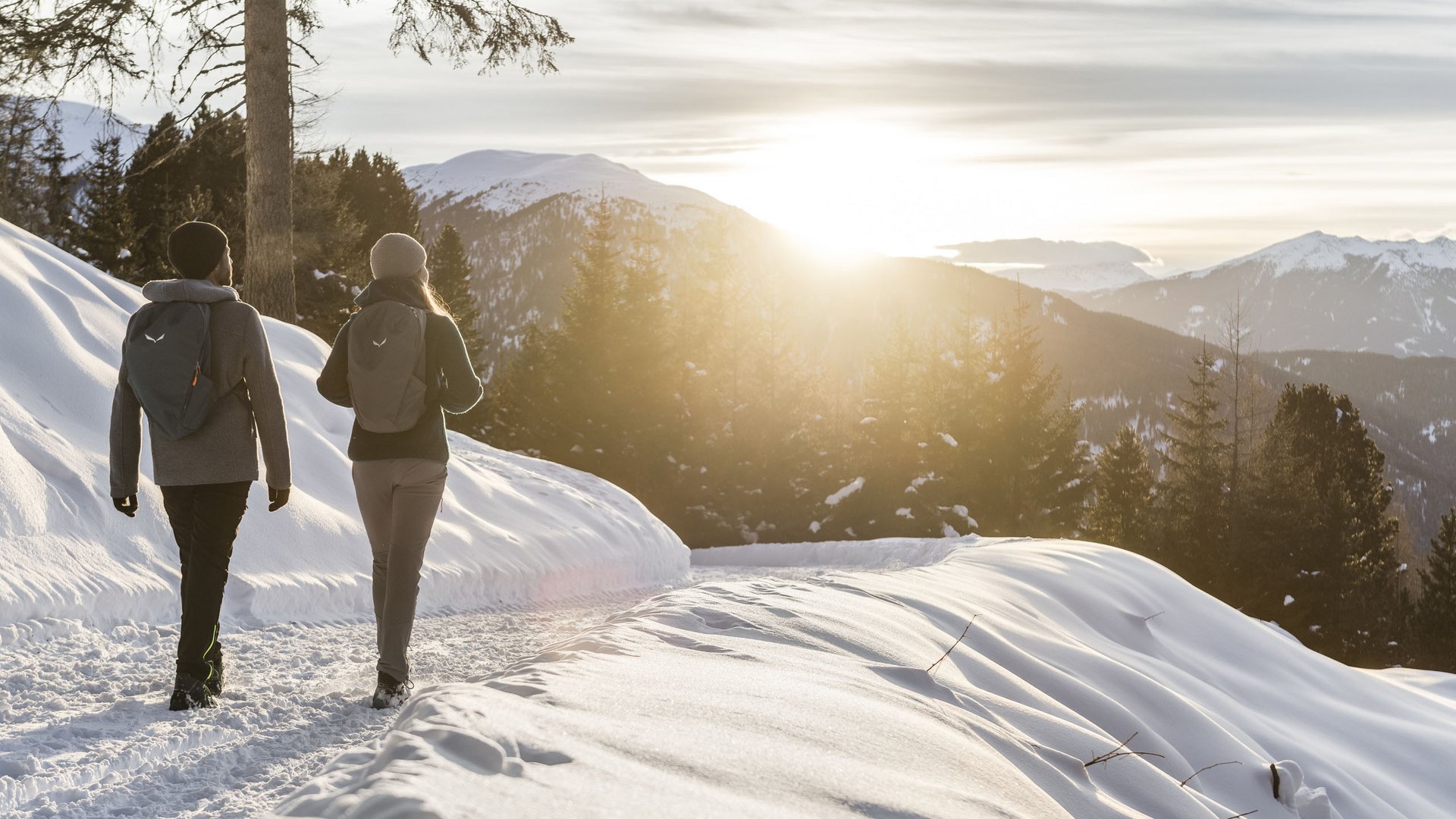 Zwei Wanderer mit Rucksäcken auf verschneitem Bergweg bei Sonnenuntergang