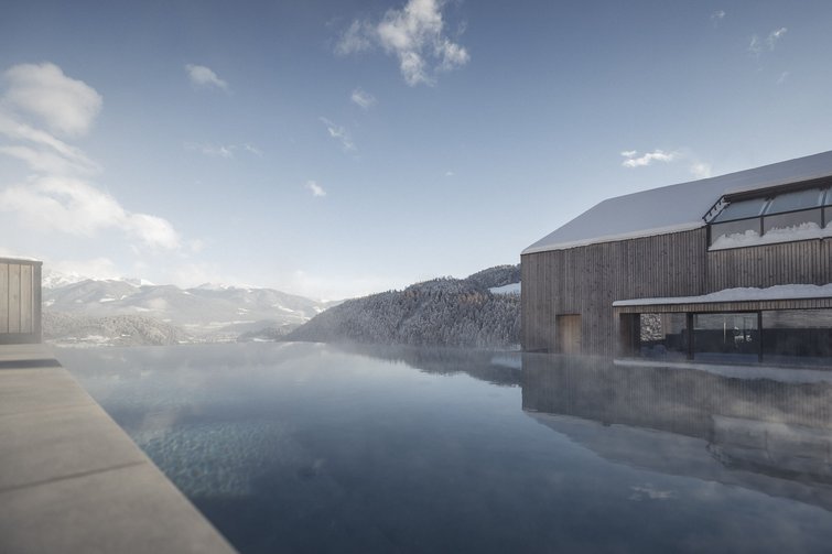 Infinity-Pool mit Bergblick und verschneitem Holzhaus unter blauem Himmel