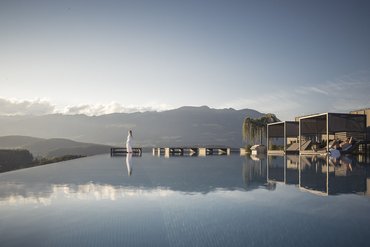 Infinity pool with mountain view, person in bathrobe and shaded lounge chairs