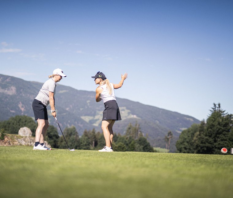 Zwei Frauen beim Golfspielen auf einem grünen Golfplatz mit Bergen im Hintergrund