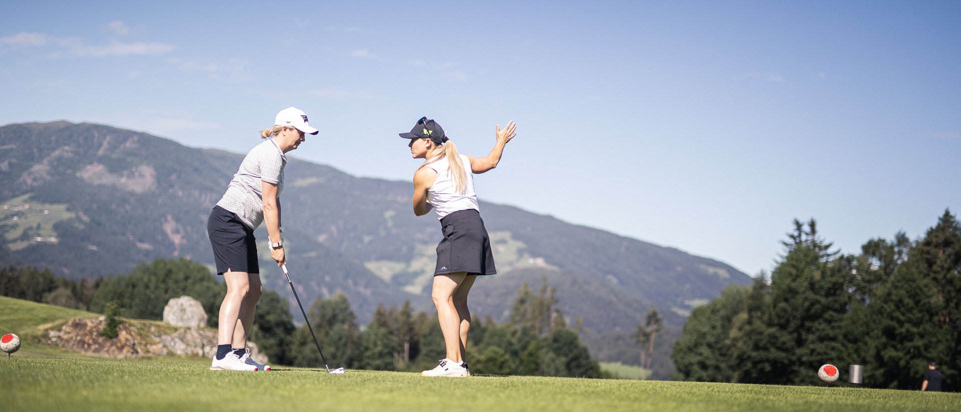 Zwei Frauen beim Golfspielen auf einem grünen Golfplatz mit Bergen im Hintergrund