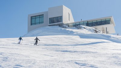 Hotel sulle piste del Plan de Corones: divertimento senza fine Due sciatori su una pista innevata davanti a edificio moderno in montagna con cielo sereno